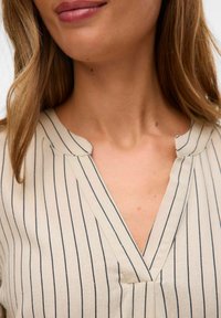 Blouse collier en beige clair avec des rayures verticales bleu marine. Dotée d'un décolleté en V et d'un tissu texturé subtil. Design minimaliste.