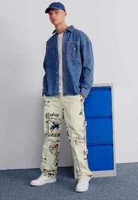 Jeans πουκάμισο με κουμπιά, λευκή μπλούζα, ανοιχτόχρωμο παντελόνι με γραφικά σχέδια, λευκά αθλητικά παπούτσια και καπέλο από τζιν. Στηρίζεται σε μια μπλε ντουλάπα.