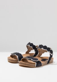 Černé semišové sandály s vlnitými pásky, zdobené kontrastními bílými akcenty. Stélka z korku a texturovaná gumová podrážka. Design s otevřenou špičkou.