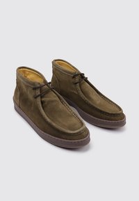 Un par de botas de ante marrón hasta el tobillo con cordones marrones oscuros y suelas de goma sobre un fondo gris claro liso.