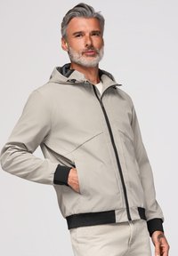 Homme aux cheveux gris et à la barbe portant une veste à capuche gris clair avec des poignets noirs et une fermeture éclair, posant avec une main dans la poche.