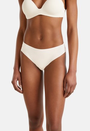 Kobieta w białym bikini pokazująca tors i górną część nóg na tle jasnego, jednolitego tła.