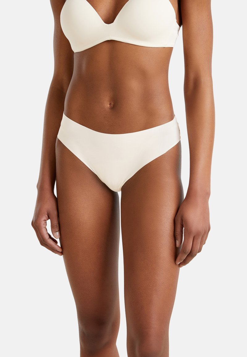 Mujer con conjunto de bikini blanco mostrando torso y parte superior de las piernas sobre un fondo claro y liso.