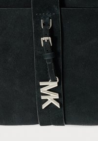 Schwarze Wildleder-Handtasche mit strukturierter Form. Sie verfügt über einen silberfarbenen "MK" Logo-Anhänger und einen glatten Ledertrageriemen mit Metallbeschlägen.