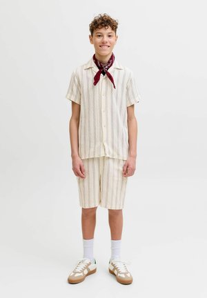 Teenager mit lockigem Haar, der ein cremefarben gestreiftes kurzärmeliges Hemd und passende Shorts, weiße Socken, Turnschuhe und ein rotes Halstuch trägt.