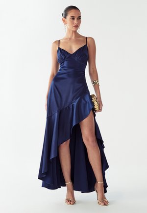 Tamsiai mėlyna satino vakarinė suknelė su aptemptu liemeniu, plonais petnešėlėmis ir aukštesne priekyje, žemesne gale, su šoniniu plyšiu; derinama su auksiniu apyrankiu ir aukštakulniais.