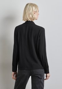 Schwarze Bluse mit langen Ärmeln aus glattem, fließendem Stoff; verfügt über einen knöpfbaren Manschetten und dezente Falten auf den Ärmeln.