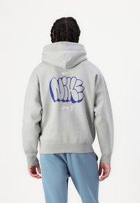 Šedá mikina s velkým modrým grafickým potiskem slova "NIKE" na zadní straně. Měkký, texturovaný materiál s uvolněným střihem a nastavitelnou kapucí.