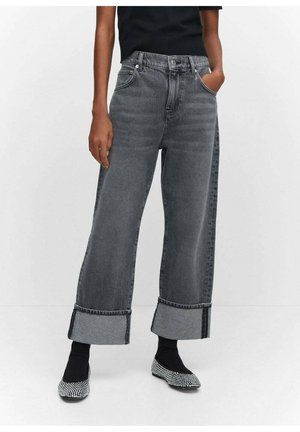 Person iført brede grå manchetjeans, sort top, sorte sokker og sort-hvide ternede flade sko, stående mod en hvid baggrund.