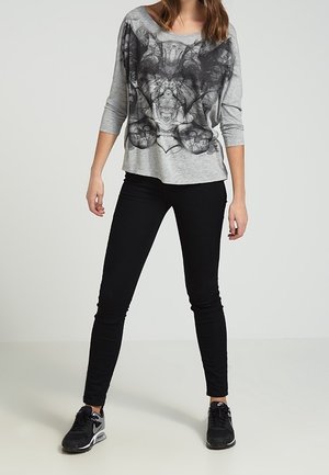 Woman wearing grey long-sleeve shirt with black abstract graphic, black fitted pants, and black Nike sneakers standing against a plain background.