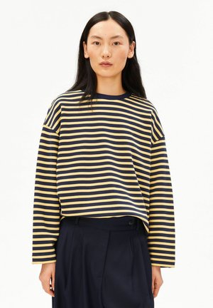 Frau mit langen schwarzen Haaren, die ein langärmeliges Shirt mit horizontalen dunkelblauen und gelben Streifen sowie dunkle dunkelblaue Hosen vor weißem Hintergrund trägt.