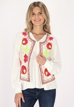 Bestickte Weste mit weißem Grund, verziert mit roten und grünen Blumenmotiven, umrandet von grauem Saum, getragen über einer hellen Bluse.