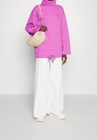 Sudadera de manga larga rosa con un alto cuello y detalle de lazo en el dobladillo, combinada con pantalones anchos blancos y un bolso suave color beige.
