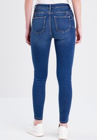 Jeans ajustados de mezclilla azul oscuro, con cintura alta, cinco bolsillos y una textura suave. Combinados con zapatillas blancas.