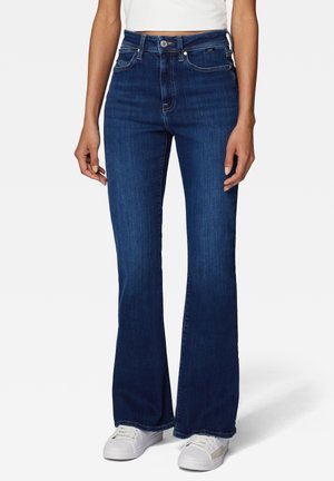 Personne portant un jean flare taille haute bleu foncé avec un bouton devant et des baskets blanches, debout devant un fond uni.