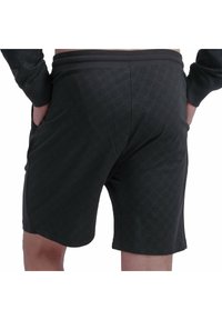 Henkilö, joka on pukeutunut mustiin kuvioituihin shortseihin, käsivarret taskuissa, kuvattu vyötäröstä alaspäin valkoista taustaa vasten.