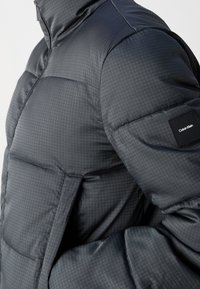 Giacca puffa in materiale grigio scuro trapuntato con un sottile motivo a quadri, presenta tasche laterali e una patch con logo sulla manica.