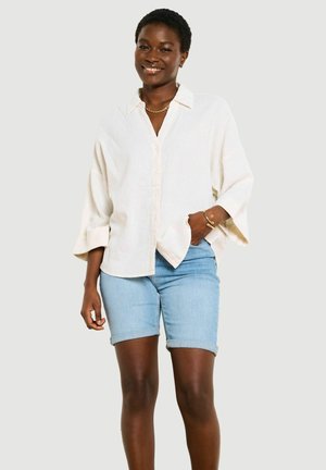 Usmievavá žena s krátkymi vlasmi, oblečená v voľnú bielu košeľu na gombíky, svetlomodré rifľové šortky, zlatý reťaz a náramky, stojaca.