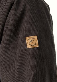 Bruine corduroy stof met fijne ribbelstructuur; voorzien van een leren patch met het label "JEANSPIRIT EST.1993" dat aan de mouw is bevestigd.