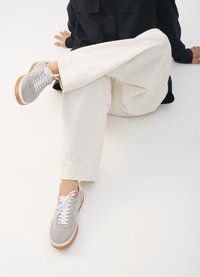 Grå semskede sneakers med hvite snørebånd og en lys brun gummisåle, sammen med løse, hvite bukser. Minimalistisk design og jevn tekstur.