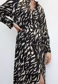 Robe chemise à motifs noirs et beiges avec de longues manches, fermeture à boutons sur le devant, une taille cintrée par une ceinture et une fente sur le côté pour faciliter le mouvement.