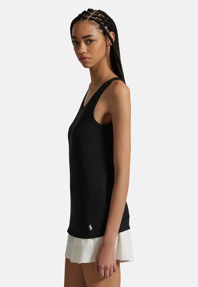 Femme aux cheveux tressés portant un haut noir sans manches et une jupe blanche plissée, debout de profil contre un fond uni.