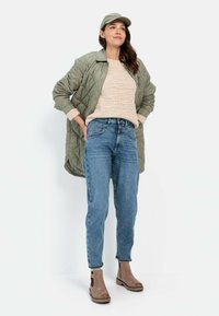 Lichtgroene gewatteerde jas, beige textuurtrui, blauwe jeans en bruine enkellaarsjes met zwarte accenten. Model poseert met handen op de heupen.