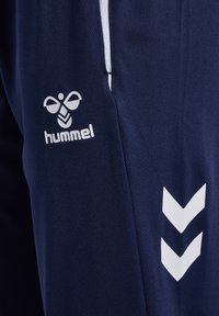 Pantalon de sport bleu marine avec logo blanc Hummel et deux bandes blanches en forme de chevrons sur la couture latérale.