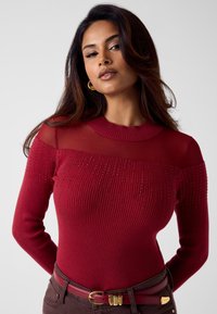 Czerwona dopasowana swetra o prążkowanej fakturze, przezroczystej górnej części, ozdobiona małymi kryształkami. Połączona z brązowymi spodniami i dopasowanym paskiem.