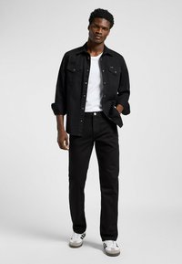 Camisa de mezclilla negra con botones al frente, combinada con jeans negros. El modelo lleva una camiseta blanca y zapatillas gris con franjas en blanco y negro.