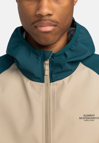 Beige jas met een teal capuchon en schouders, ritssluiting en logodetails. Kenmerkt zich door een soepele textuur en een sportief, functioneel ontwerp.