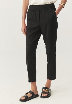 Persoană îmbrăcată cu pantaloni scurți negri, cu talie elastică și sandale negre de tip slip-on, cu detalii aurii, stând pe un fundal alb.