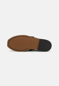 Schwarze Leder-Schlüpfschuhe mit einer braunen Gummisohle, dekorativen Schnallenakzenten und kontrastierenden Nähten entlang des Randes.
