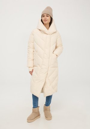 Cappotto imbottito crema, lungo fino al ginocchio, con motivo a rombi diagonali, cappuccio oversize e tasche laterali. Abbinato a jeans azzurri e stivali beige.