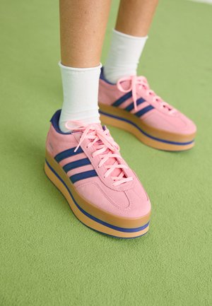Lyserøde ruskinds platform sneakers med blå striber og lyserøde snørebånd. Tyk tan sål med blå accent. Båret med hvide ankelstrømper på en grøn overflade.
