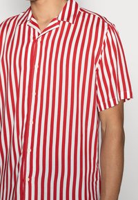 Camisa de manga corta con rayas verticales rojas y blancas, tela ligera, cuello con muesca y cierre frontal con botones.