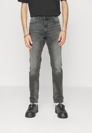 Mand står iført falmede sorte slim-fit jeans, sorte sneakers, hvide sokker, sort t-shirt og guldfarvede og sorte armbånd på håndleddet mod en hvid baggrund.