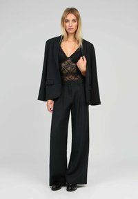 Femme portant un haut en dentelle noire, un blazer noir surdimensionné posé sur les épaules, un pantalon large noir et des chaussures noires, se tenant contre un fond gris.