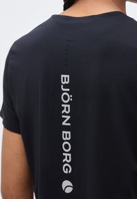 Άτομο που φοράει μαύρο πουκάμισο με την επιγραφή "BJÖRN BORG" τυπωμένη κατακόρυφα σε λευκό χρώμα στο πάνω μέρος της πλάτης, κοντά στον λαιμό.