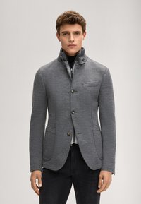 Szara kurtka z teksturowanej tkaniny, z wysokim kołnierzem, czterema guzikami oraz dwoma przednimi kieszeniami. Nosi się ją na czarnym golfie.