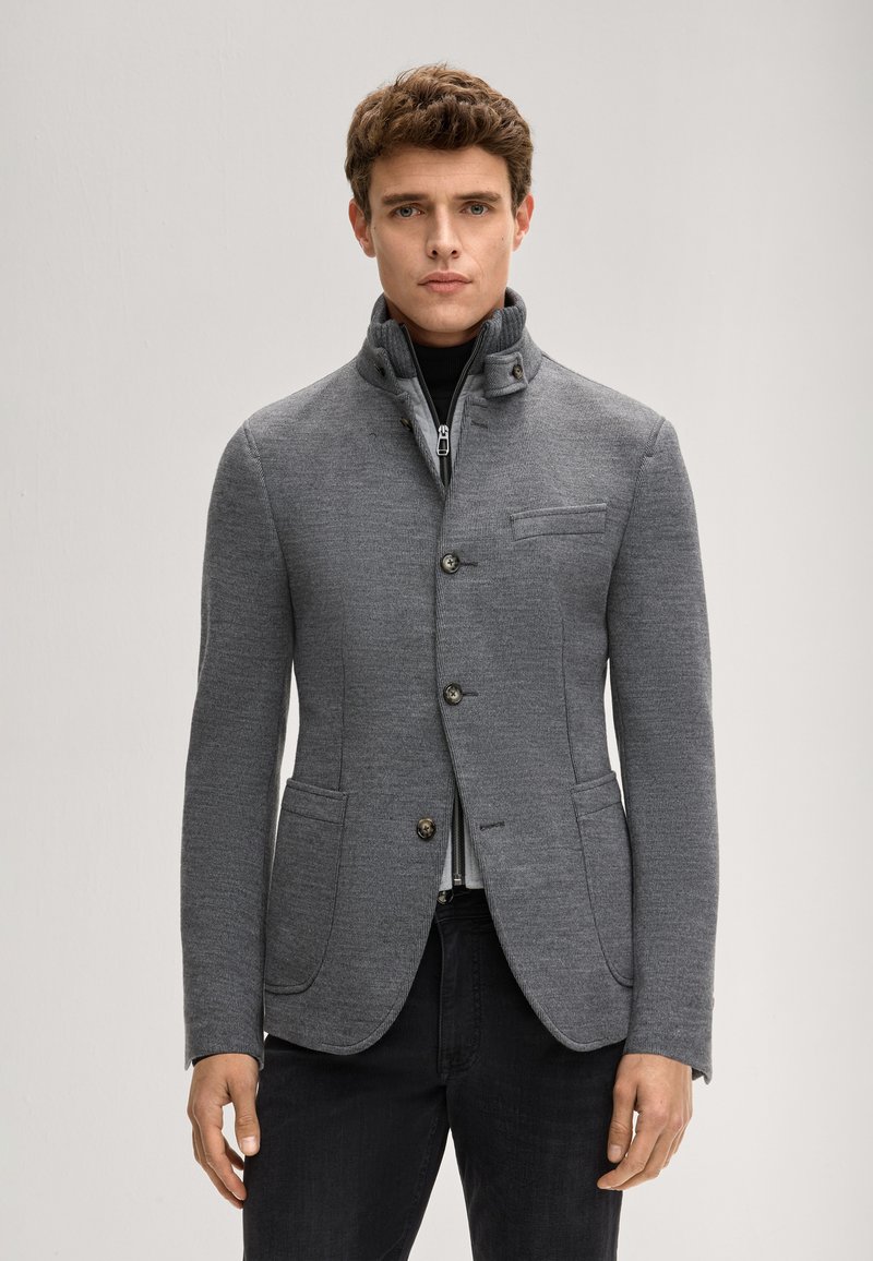 Szara kurtka z teksturowanej tkaniny, z wysokim kołnierzem, czterema guzikami oraz dwoma przednimi kieszeniami. Nosi się ją na czarnym golfie.