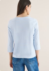 Femme aux cheveux longs et bruns portant une chemise à manches trois-quarts rayée bleu clair et blanc et un jean bleu, tournée dos à la caméra.