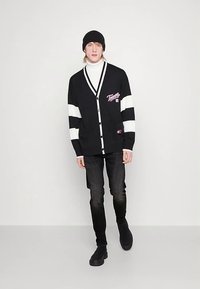 Black cardigan with white stripes and a front pocket, buttoned design, worn over a white turtleneck, paired with dark jeans and black shoes.