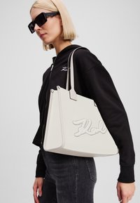 Balta-offa somiņa ar teksturētu materiālu, trīsstūra formu un ieplānoto "Karl" logotipu. Aprīkota ar diviem augšējiem rokturiem un gludu apdari.