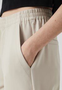Beige bukser med elastisk talje, sidelomme og glat, tekstureret stof. Nærbillede af en hånd hviler i lommen.