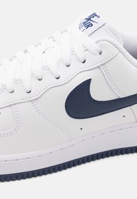 Bijele kožne tenisice s tamnoplavim akcentima, koje imaju istaknuti Nikeov swoosh logo, teksturiranu površinu i ravne bijele vezice.