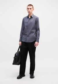 Γκρι πουκάμισο με υφή απαλή, συνδυασμένο με μαύρο παντελόνι. Το μοντέλο κρατάει μια μαύρη δερμάτινη τσάντα και φοράει μαύρα παπούτσια.