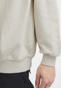 Sudadera color beige con puños y dobladillo acanalados. Textura visible; costura lateral con pequeño detalle de etiqueta. Mano posicionada cerca del lado inferior.