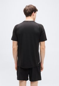 Μαύρο αθλητικό t-shirt με κοντά μανίκια, λεία υφή, καμπυλωτό στρίφωμα και αντανακλαστικές λεπτομέρειες στην πλάτη. Φορεμένο με αντίστοιχα σορτς.