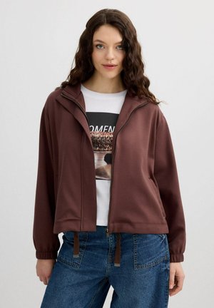 Mujer con cabello castaño ondulado lleva una chaqueta marrón con cremallera sobre una camiseta gráfica blanca y jeans azules, de pie frente a un fondo claro y liso.